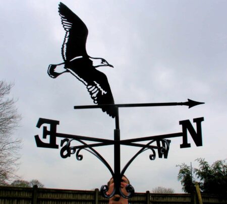 black outline Flying Albatross Bird Weathervane against a blue-grey sky and dark hedgerow