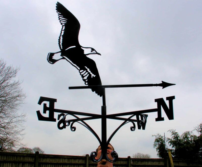 black outline Flying Albatross Bird Weathervane against a blue-grey sky and dark hedgerow