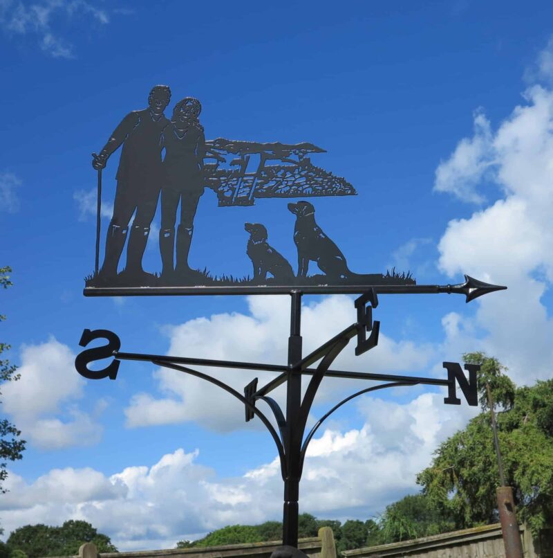 a dog walker themed weathervane - picturing a Couple Walking, Hills And Dogs against a blue sky